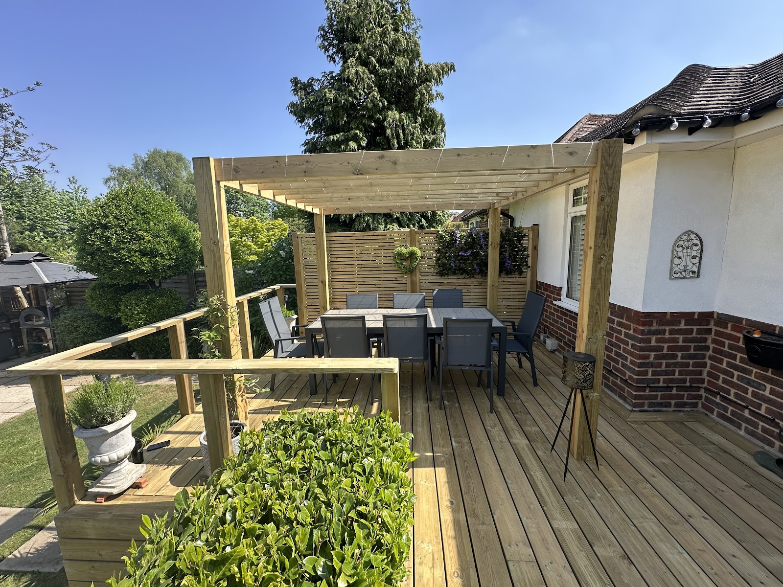 A raised timber deck with a pergola and dining set, built out to catch the afternoon sun over a mature planted boundary.