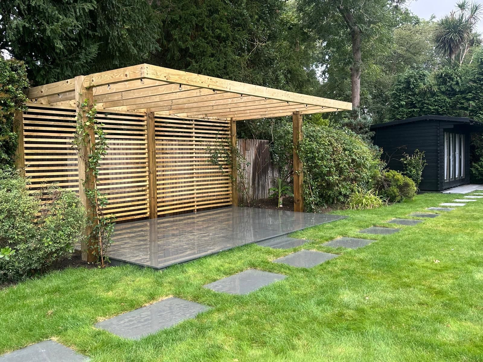 A contemporary slatted pergola over a porcelain-paved sitting area, with slate stepping stones set through the lawn to reach it.